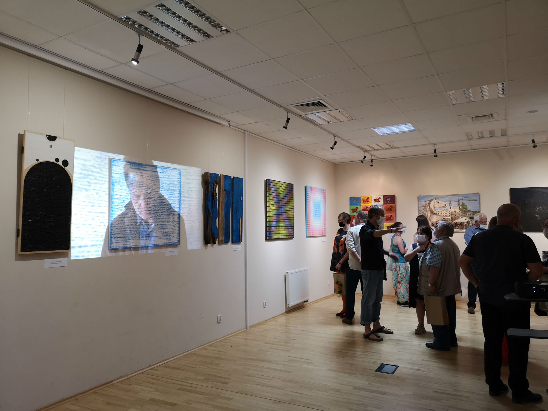 Fotografia wykonana w sali konferencyjno-wystawowej w Miejskiej Bibliotece Publicznej w Mińsku Mazowieckim. Z lewej strony ściana z zawieszonymi obrazami, między którymi wyświetlany jest film z rzutnika. Prawą stronę kadru wypełniają zwiedzający.