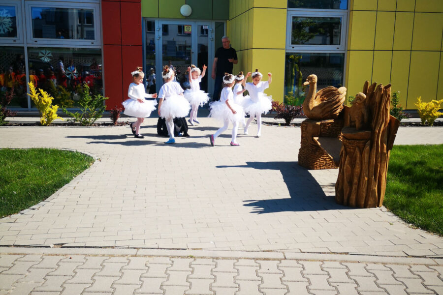 Fotografia wykonana przed przedszkolem miejskim nr 5. Dziewczynki, przebrane w stroje białe stroje baletnic, wykonują taniec. Z prawej strony kadru stoi drewniana ławka dekorowana rzeźbiarsko motywami zwierzęcymi.