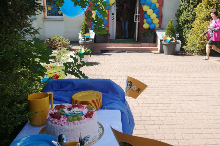 Fotografia wykonana przed przedszkolem miejskim nr 3. Na pierwszym planie stół z tortem. W tle wejście do przedszkola. Wszystko udekorowane żółto-niebieskimi balonami oraz chorągiewkami przypominającymi o obchodach 600-lecia miasta.
