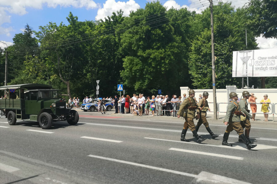 Parada z okazji obchodów 100-lecia przybycia 7 Pułku Ułanów Lubelskich do Mińska Mazowieckiego. Ulicą miasta idzie czterech żołnierzy ubranych w historyczne, zielone mundury; na głowach mają hełmy. Za nimi podąża zabytkowy samochód wojskowy.
